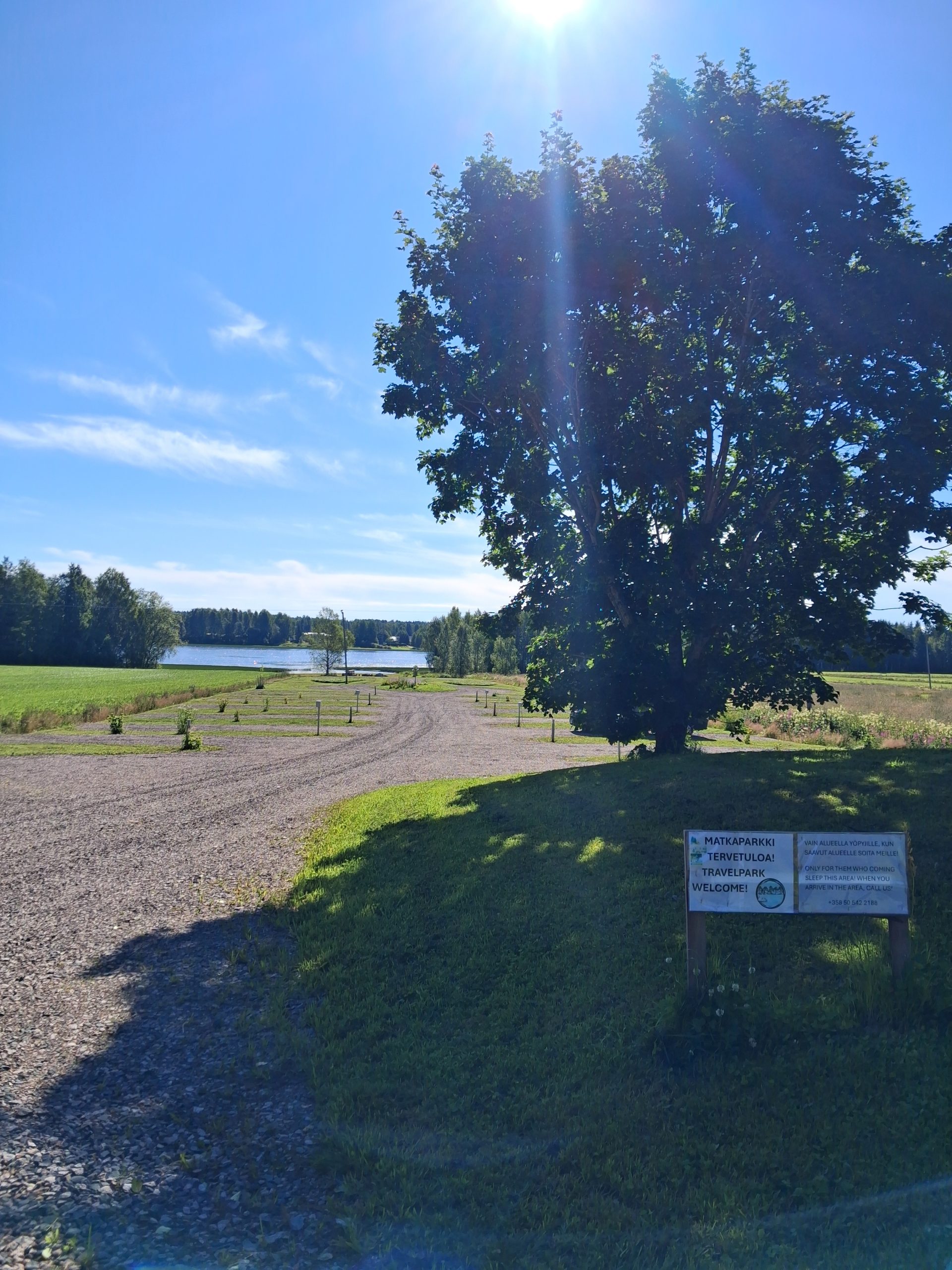 Sisääntulo matkaparkkiin ja sähköpaikat Aurinkoinen päivä ja aurinko paistaa puun takaa, sekä sähkötolppia maassa.